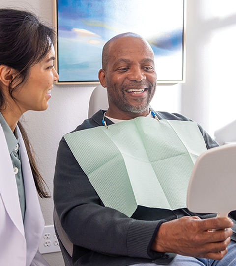 A medical professional in a white coat looks on as a patient smiles at the reflection in a handheld mirror.