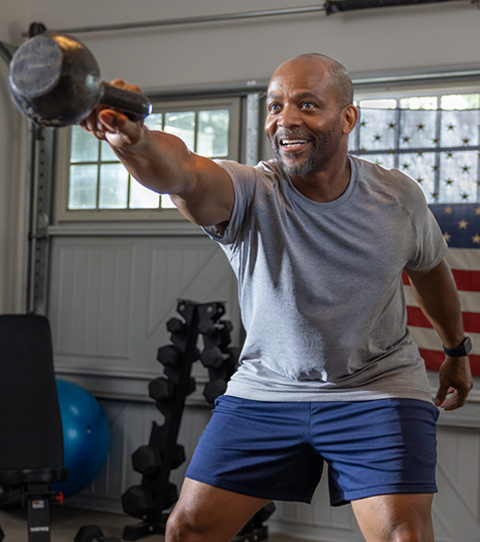 A person in workout clothes is in a garage, lifting a kettlebell with one arm and looking strong.