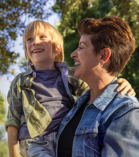 A parent in a denim jacket holds a young child in an outdoor setting; both are smiling and looking upward.
