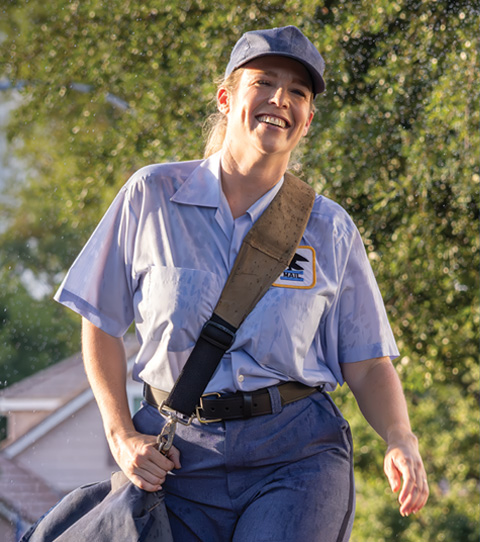 A happy mail carrier in uniform smiles on the job, with sprinklers watering the lawn.